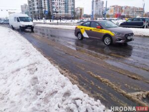 Слякотная улица с заснеженными тротуарами, припаркованный фургон и проезжающее такси под облачным небом.